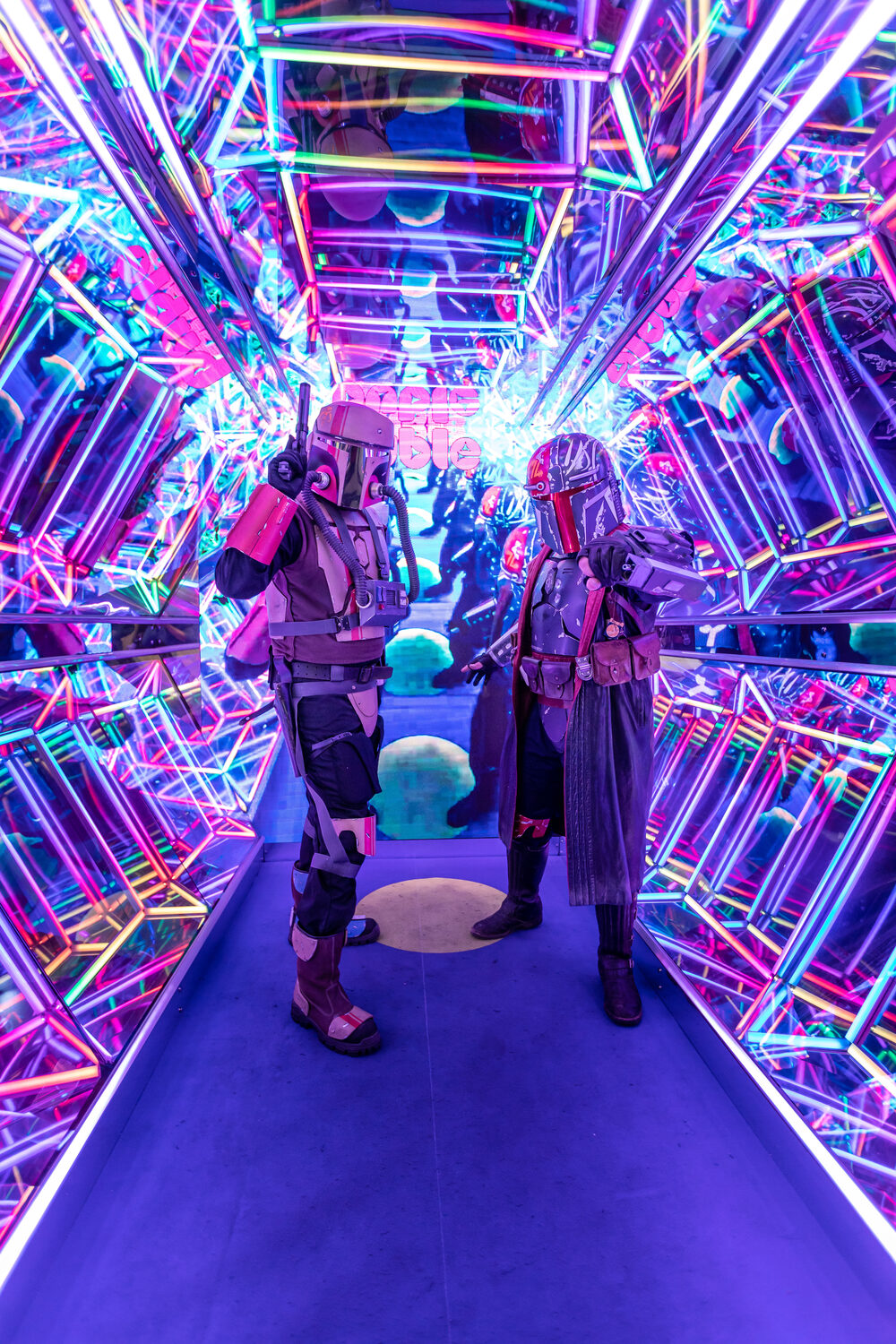 Two costumed visitors standing inside the Infinity Cave — a mirrored corridor of multicoloured LED lattices repeating away to infinity around them.