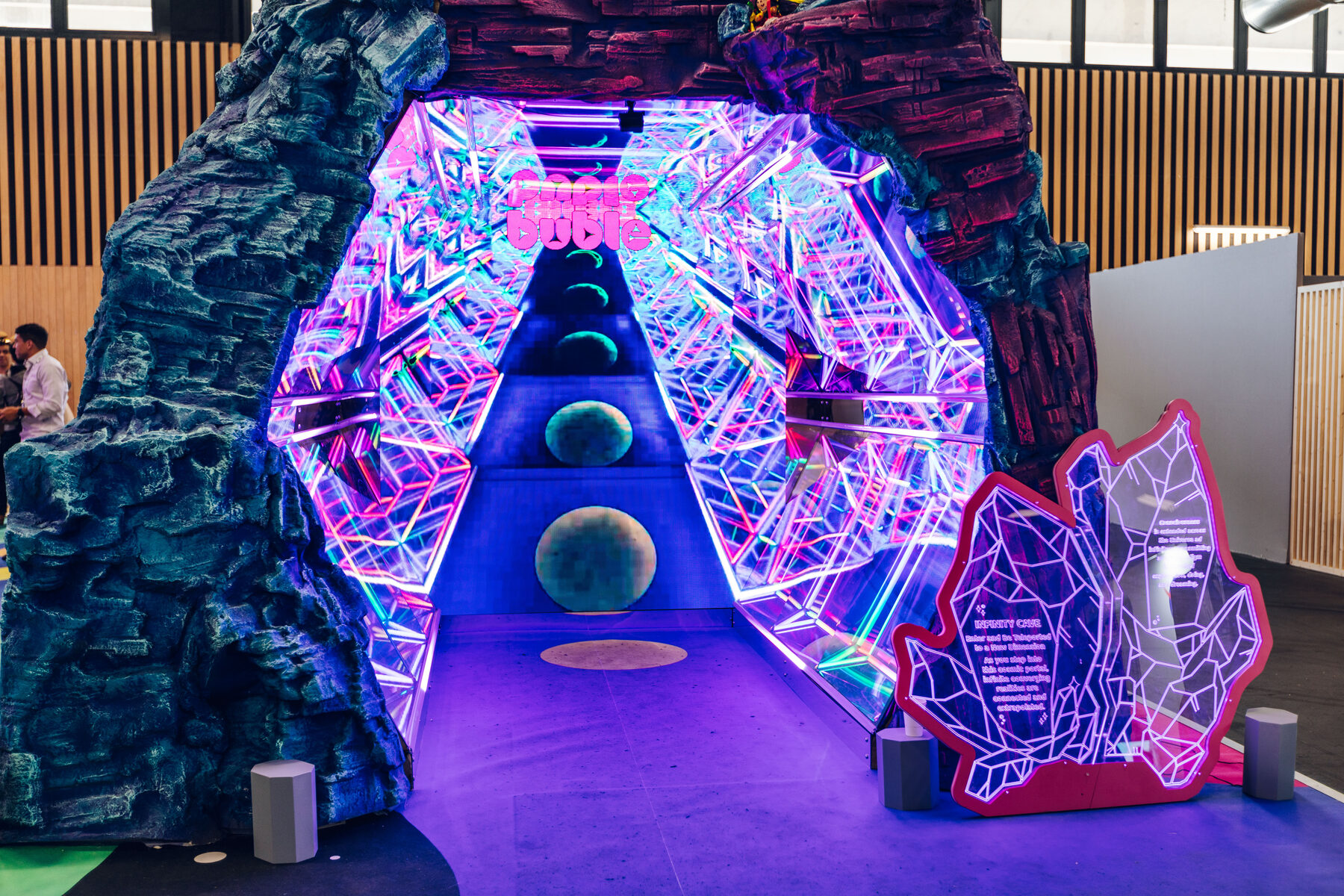 The Infinity Cave from outside — a dark rocky portal opening onto a mirrored interior of endless multicoloured LED lattices, with green stepping stones leading in.