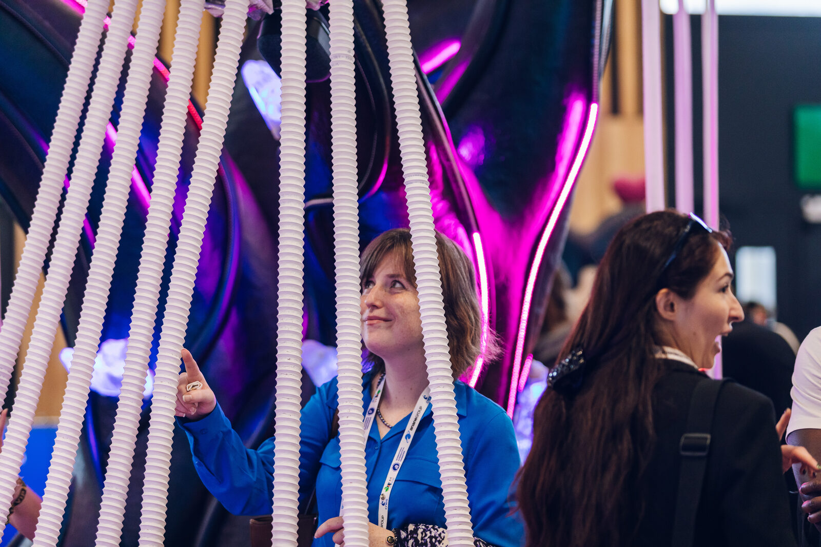 Two visitors standing inside the fan of ribbed white musical tendrils running from the canopy to the floor, one reaching up to play them, the glowing purple trunk behind.
