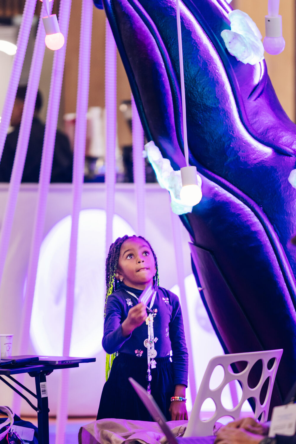 A young girl reaching up to touch one of the tree's hanging light-ball tendrils, lit softly beneath the canopy.