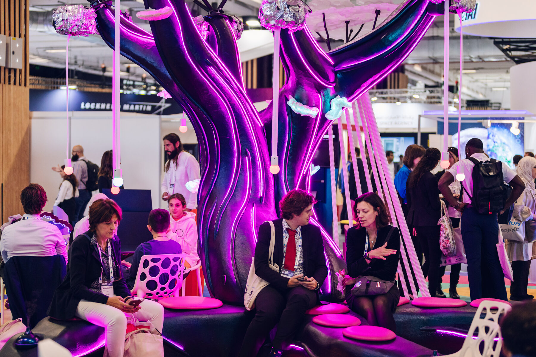 Delegates sitting and talking on the seating beneath the alien tree's glowing canopy, tendrils hanging around them.