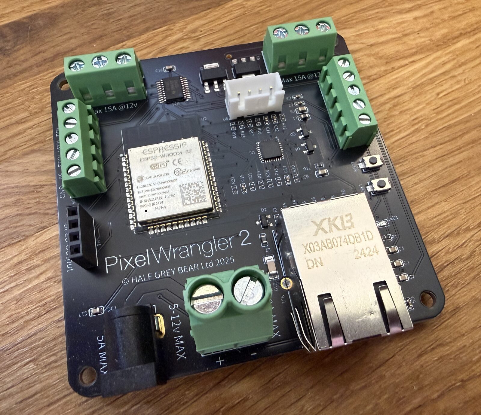 A custom black circuit board labelled PixelWrangler 2, © HALF GREY BEAR Ltd 2025, with an ESP32 module, Ethernet jack, screw terminals and DC power input.