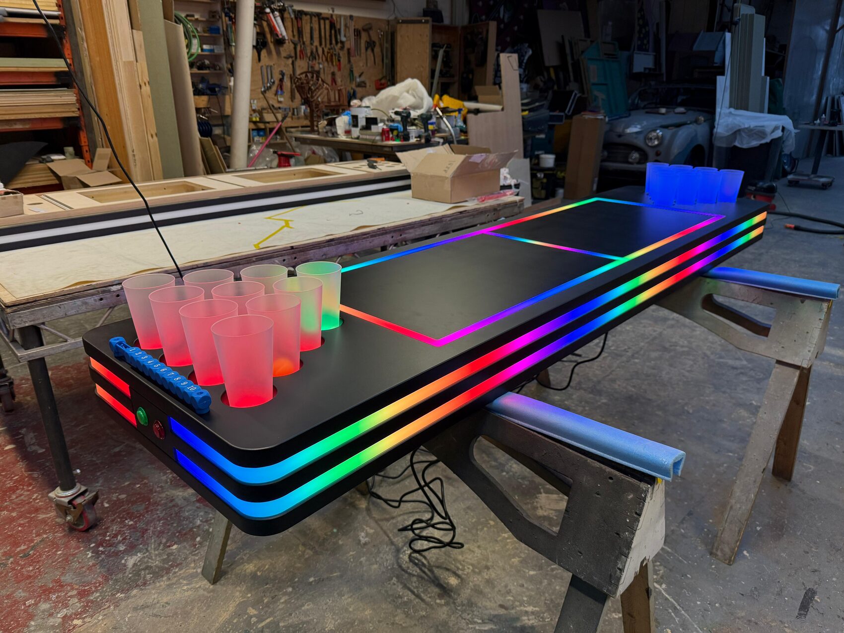 Both tables in the workshop with their cup racks lit — one end glowing red, the other blue — and rainbow pixel tape running around the table edges.