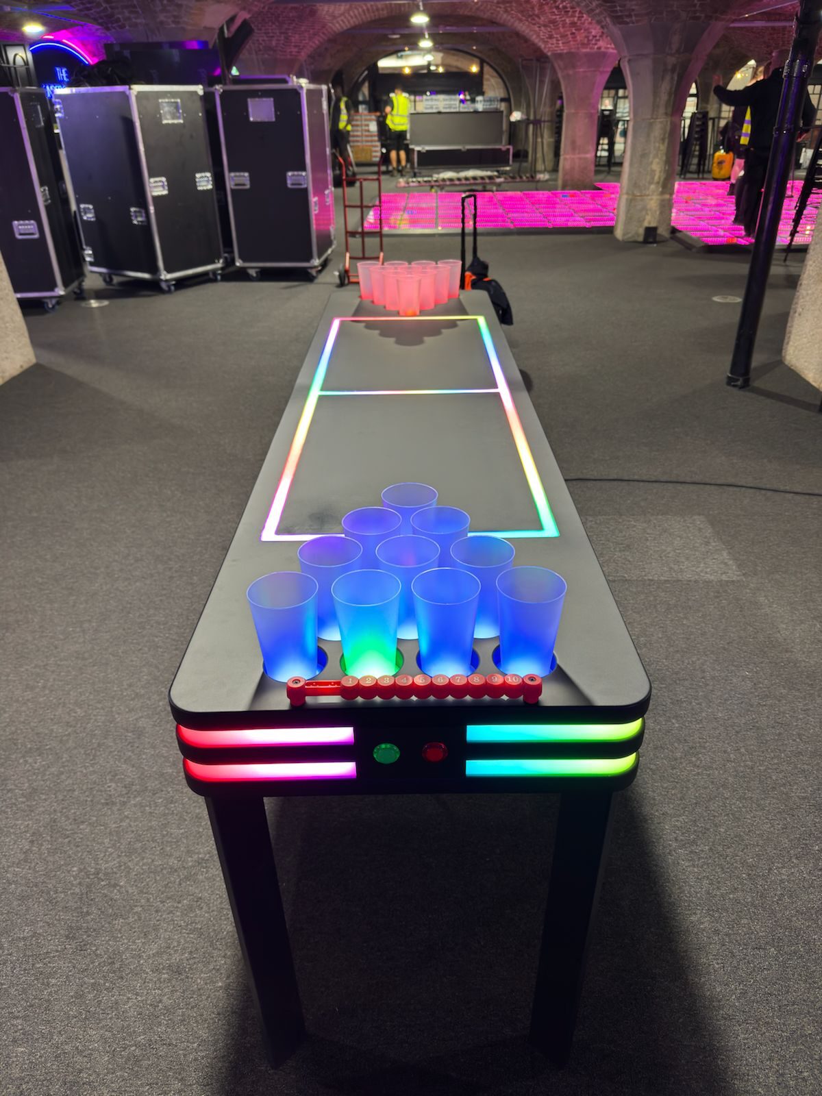 A finished LED neon beer-pong table on site under the brick vaults of Tobacco Dock — rainbow pixel-tape edges glowing, blue-lit cups racked at the near end.