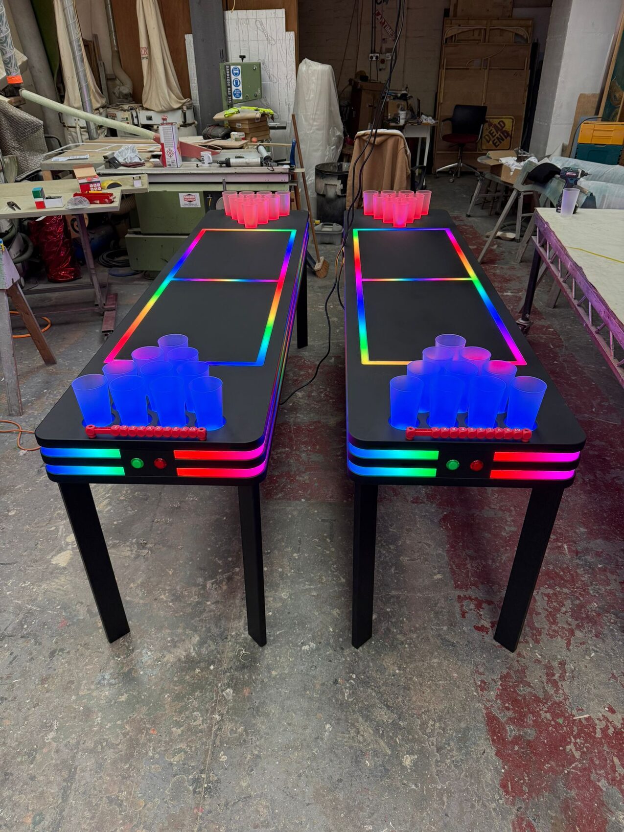 Two LED neon beer-pong tables side by side, cup racks glowing red and blue, rainbow pixel-tape edges.