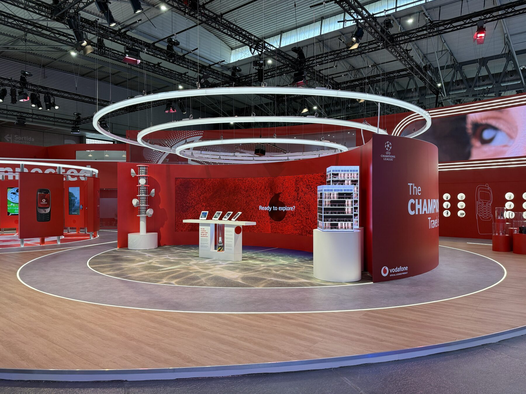 A clean wide view of the Vodafone stand at MWC 2026 — a comms-tower prop, iPad interactive plinths and a subsea-cable display under concentric ring lights, with Champions League branding.