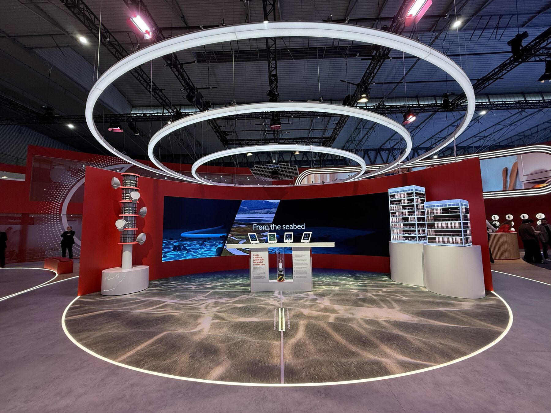 The Seabed to the Stars installation — three concentric light rings over the iPad console and undersea-cable model, the curved LED wall playing 'From the seabed', the cell tower to the left, the LED building towers to the right, and caustic water light rippling across the floor.